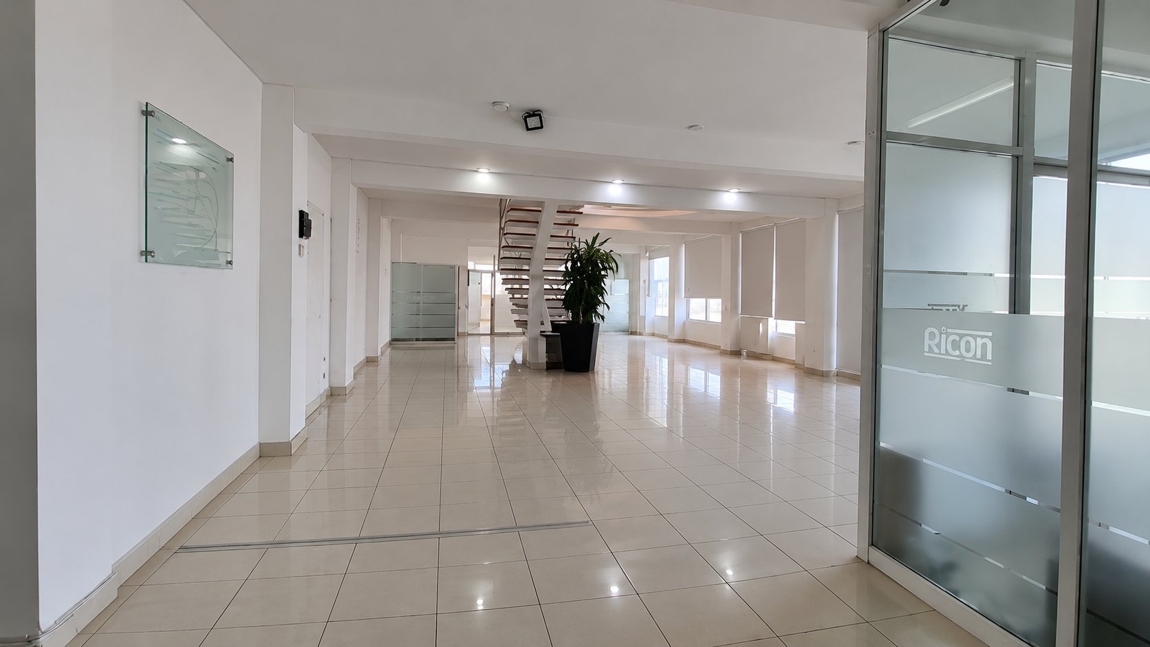 Planta abierta con piso de cerámica, escalera blanca al fondo y cancel de vidrio esmerilado con el logotipo de Ricon
